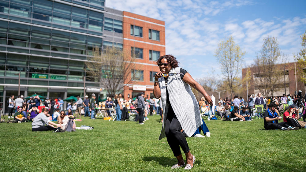 A person dances while holding solar eclipse glasses. They are standing on Merten Lawn, where many people are gathered to view the solar eclipse.