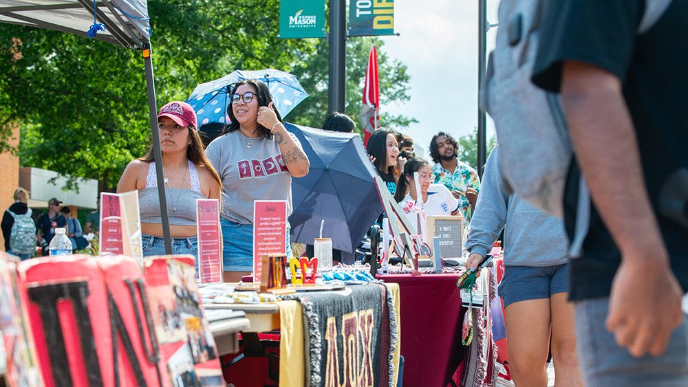 Students walk by booths for different Greek Life organizations. The people staffing the booths are engaging with different students.